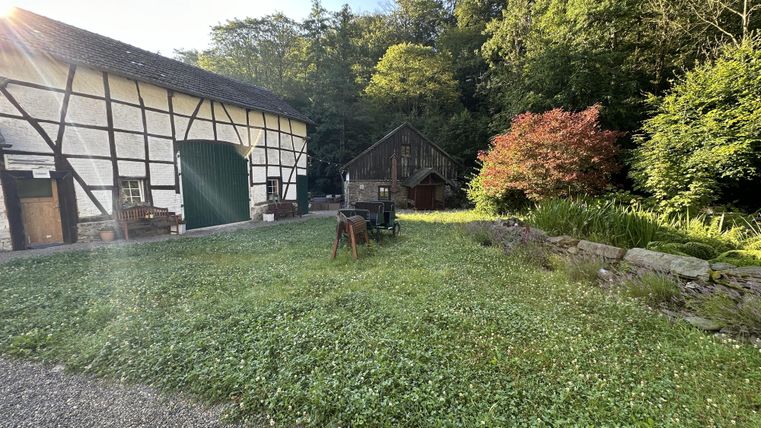 Ein idyllischer Hof mit einem alten Fachwerkhaus und einer gemütlichen Scheune. Der grüne Garten ist umgeben von Bäumen und Blumen.
