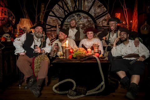Een groep piraten zit om een tafel met voedsel en dranken. De sfeer is feestelijk en de omgeving is in piratenstijl gedecoreerd.