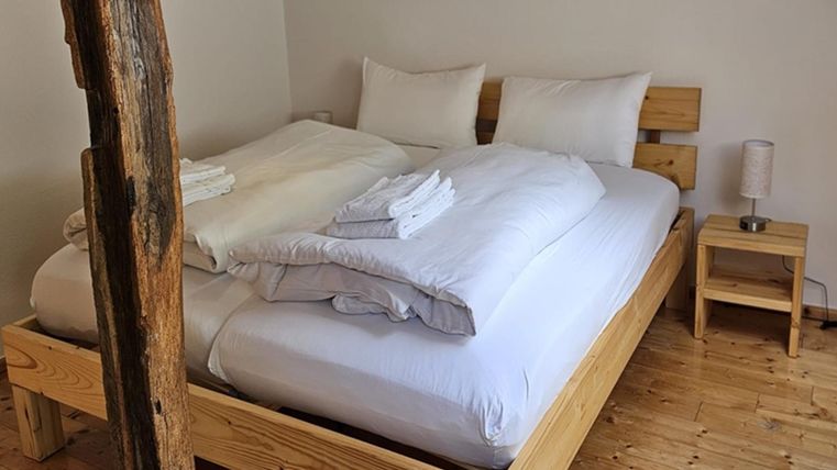 Ein gemütliches Schlafzimmer mit einem Holzbett und frohen weißen Bettwäsche. Neben dem Bett steht ein kleiner Nachttisch mit einer Lampe.